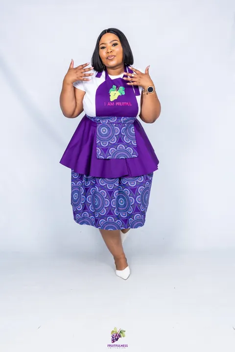 A smiling woman in a purple I AM FRUITFUL apron with a mandala-print pocket and matching skirt
