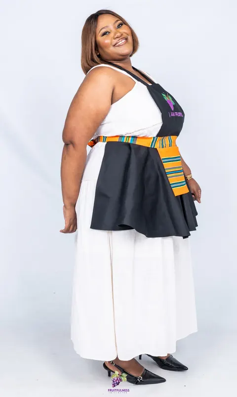 Side view of a smiling woman in a black apron with a striped orange Munwenda pocket, worn over a white dress