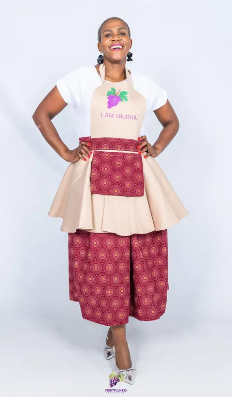 A woman laughing in a beige I AM FRUITFUL apron with a maroon-print pocket and skirt, hands on her hips